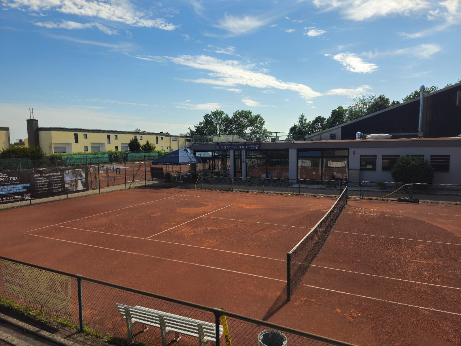 Roter Tennisplatz mit Netz vor dem Gebäude des TSG Sportcenters.