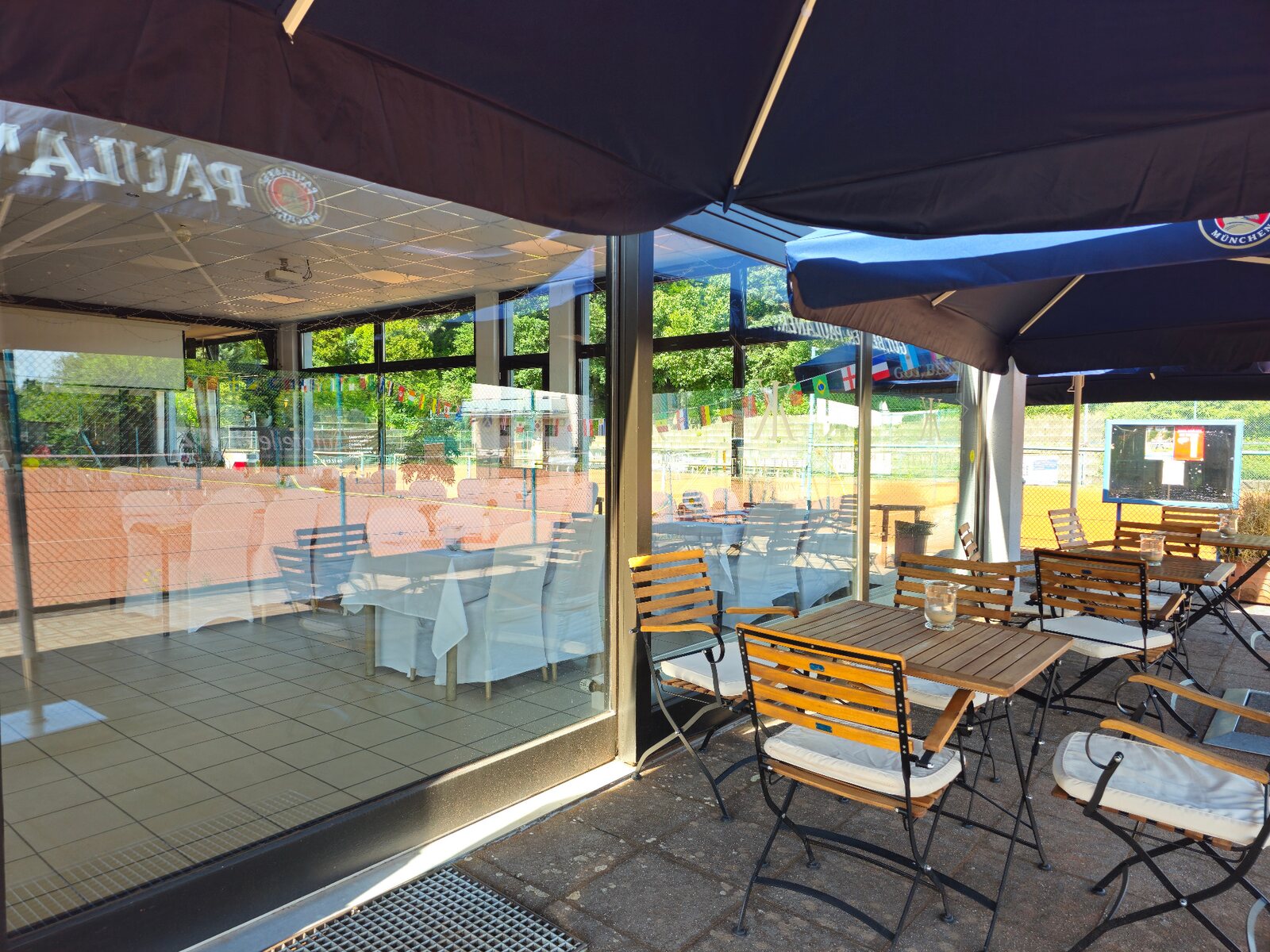 Holztische und Stühle unter Sonnenschirmen neben der Glasfront des Clubgebäudes.